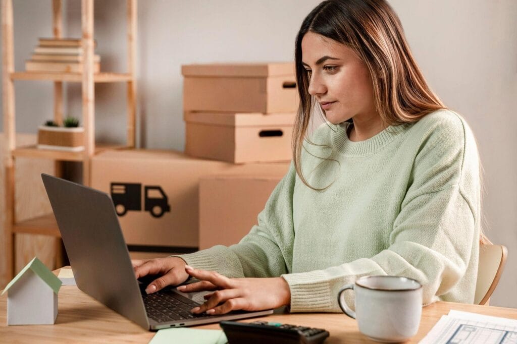 A young woman in a home office working on a laptop with shipping boxes in the background, representing an e-commerce business.